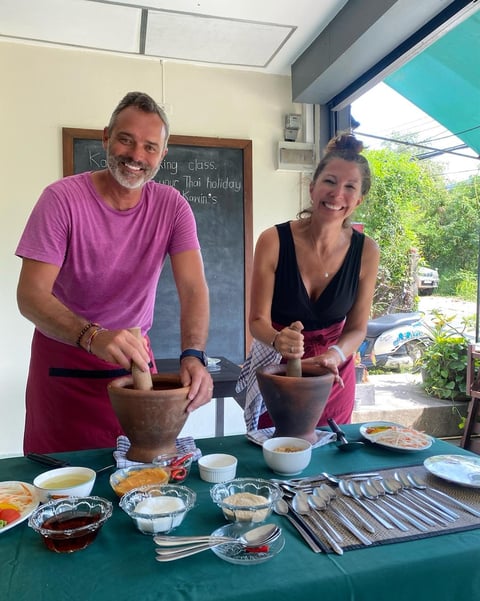 Two people in a cooking class demo, both using large wooden mortars and pestles, with ingredients and utensils on a green table in front of them