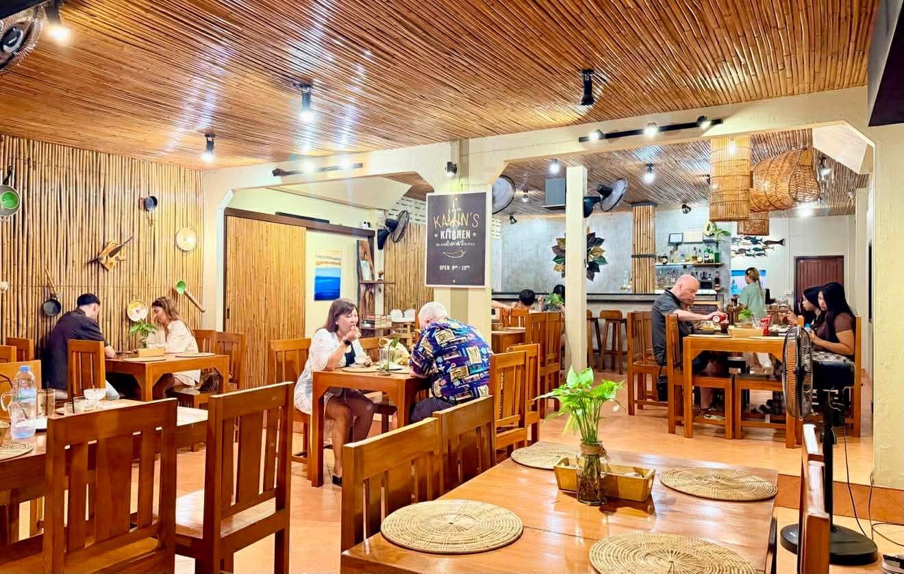 Warm wooden cafe interior with customers seated at tables, featuring wooden ceiling, furniture, and potted plants
