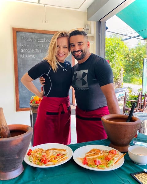 Two people in matching red aprons smile in a bright kitchen with cooking class dishes on a turquoise counter
