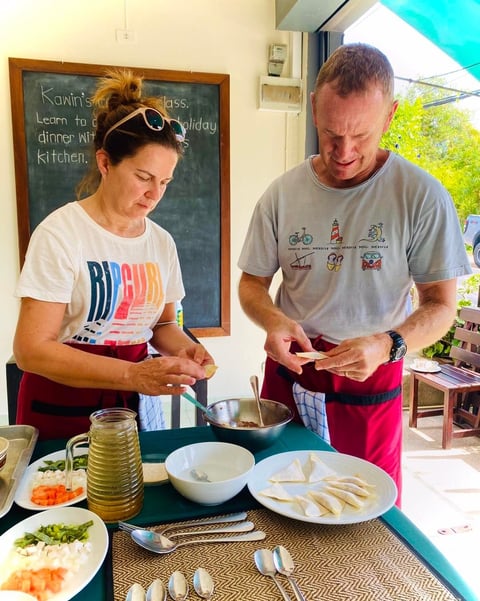 Two people cooking together at an outdoor kitchen counter with fresh ingredients and utensils visible on the table