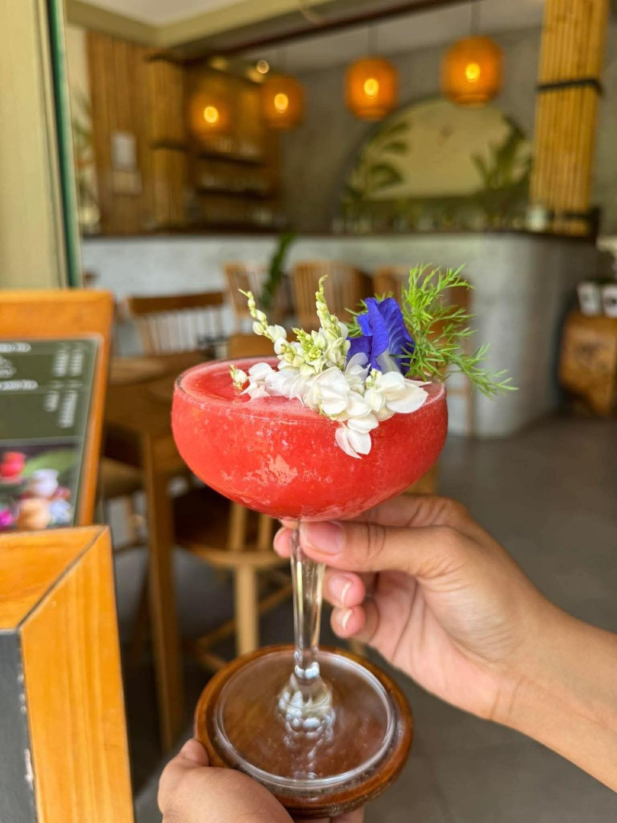 Hand holding a vibrant red cocktail garnished with white flowers, blue iris, and fresh herbs in a modern bar setting