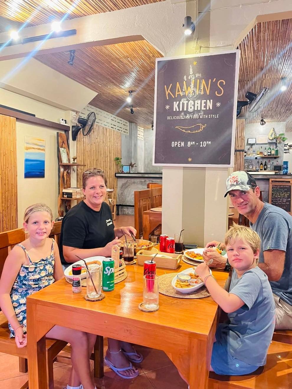 Four people dining at a wooden table in Kawins Kitchen restaurant with decorative wood ceiling and signage visible