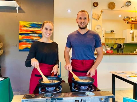 Two people wearing red aprons cooking together on stovetops in a bright kitchen with colorful artwork on walls