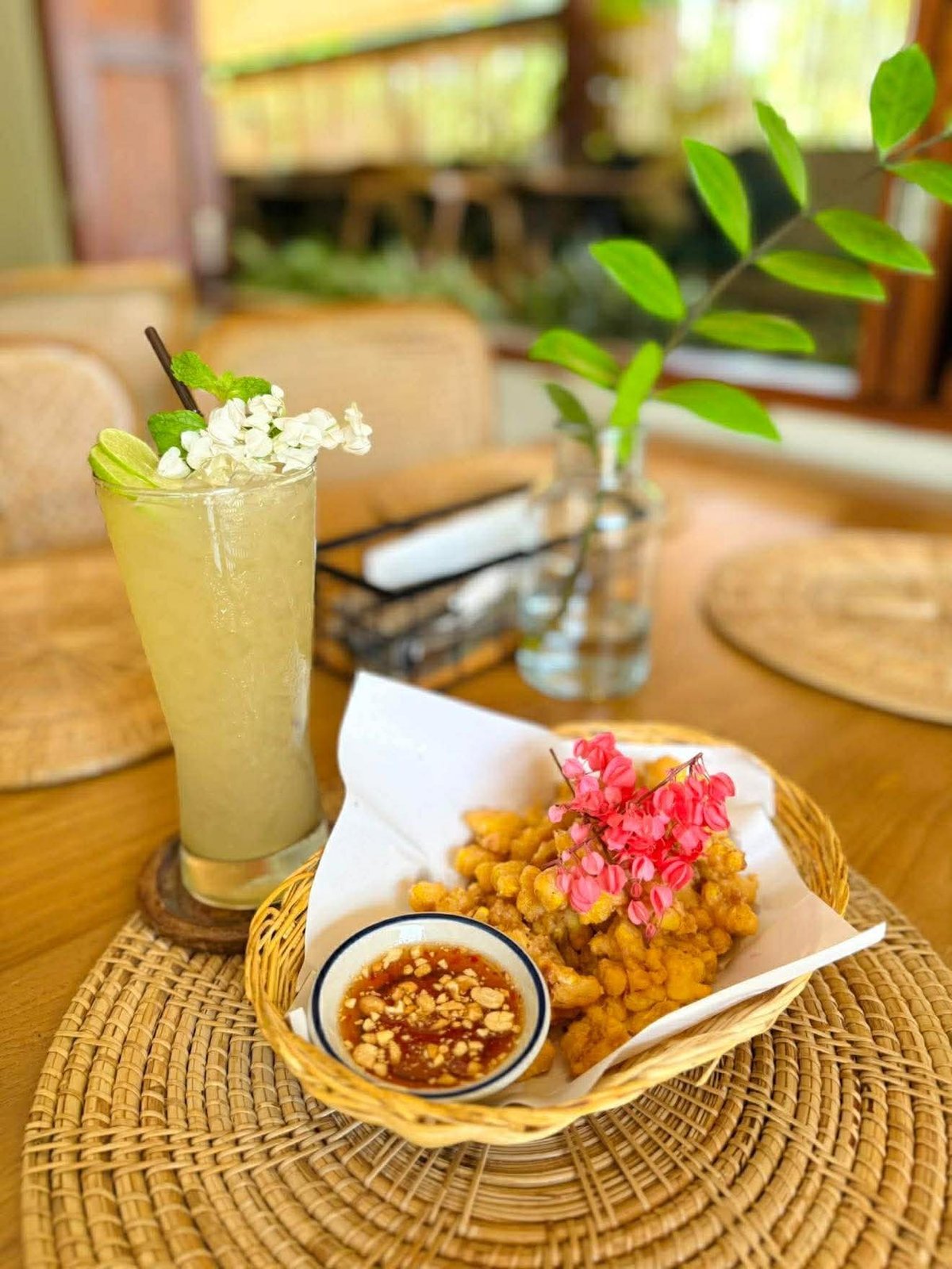 Refreshing citrus beverage with mint and white flowers next to golden fried appetizers with pink flower garnish and dipping sauce on woven placemat