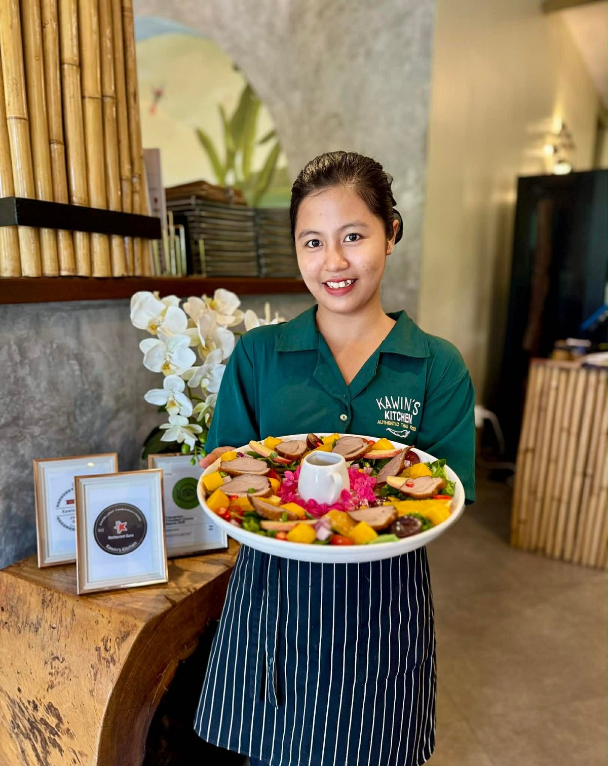 Woman in green chefs uniform and striped apron holding a colorful fruit and vegetable platter in a modern restaurant setting