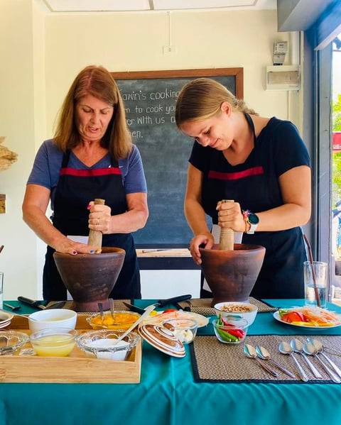 Two women in aprons using mortars and pestles at a cooking class counter with ingredients and bowls arranged in front