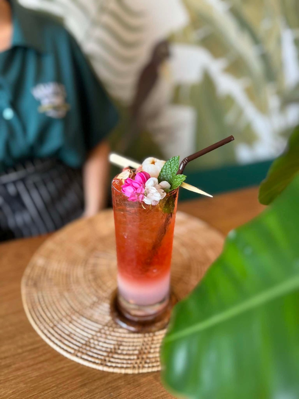 Tropical cocktail with red liquid, ice, and pink flower garnish on wooden coaster, held by server in teal uniform
