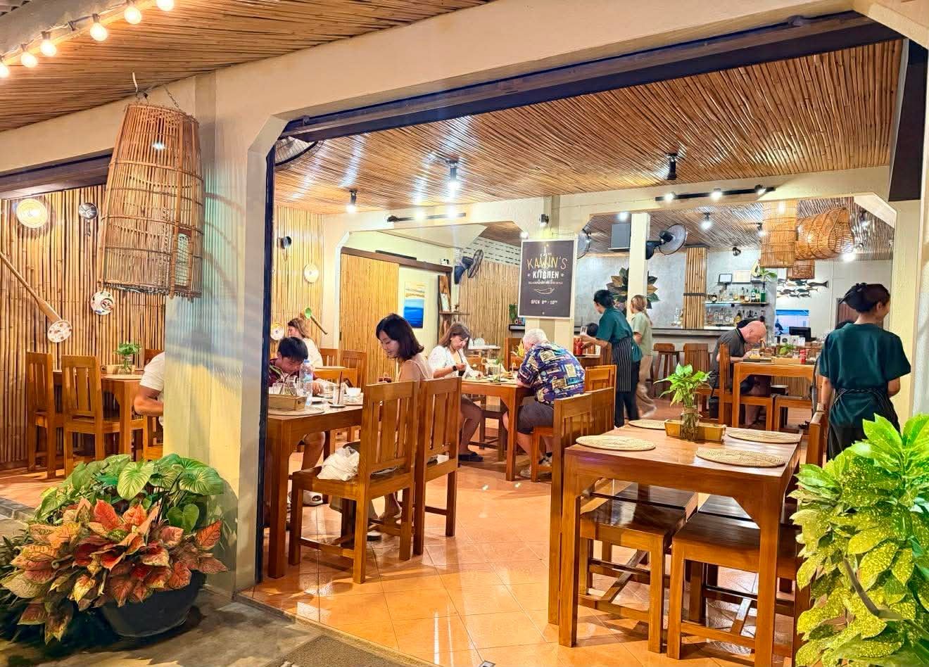 Warm, wooden interior of a casual restaurant with dining tables, woven pendant lights, and potted plants throughout the space