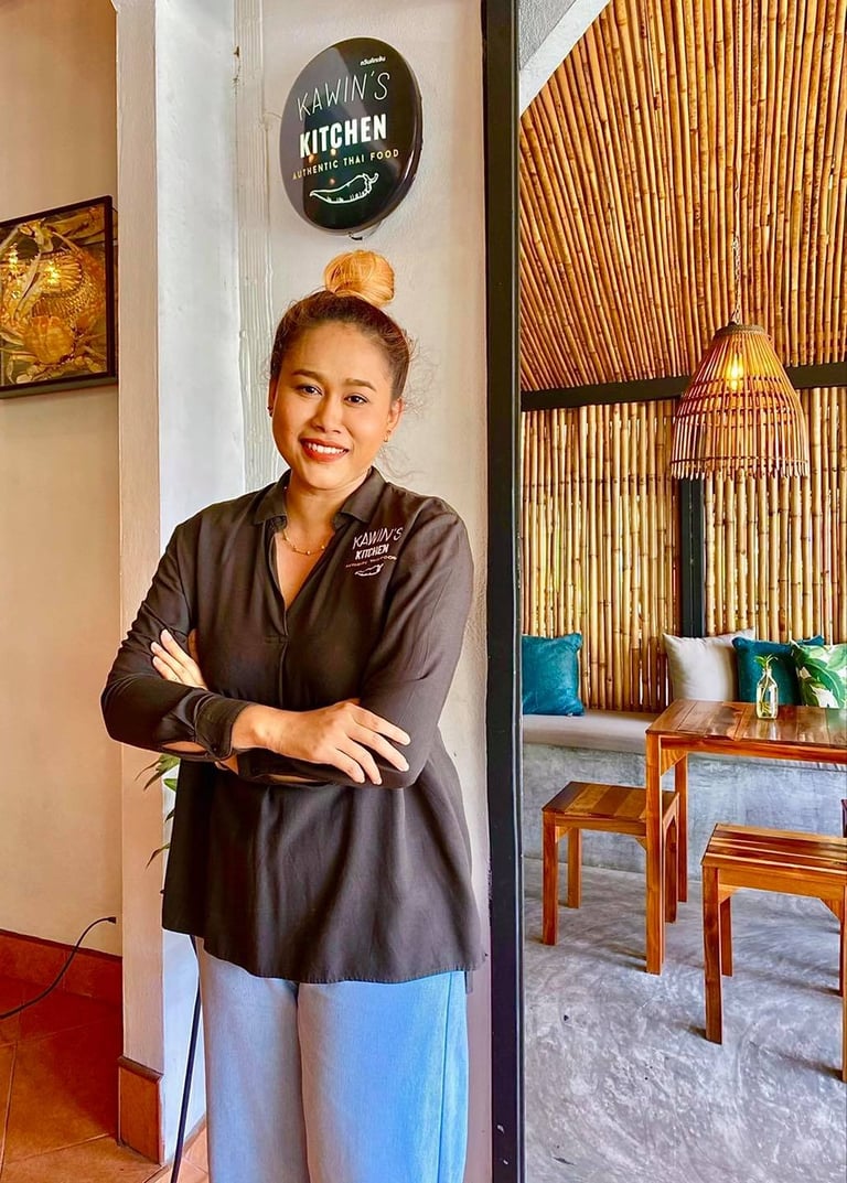 Woman in black chef coat standing in restaurant entrance with arms crossed, smiling at camera near Ramins Kitchen sign