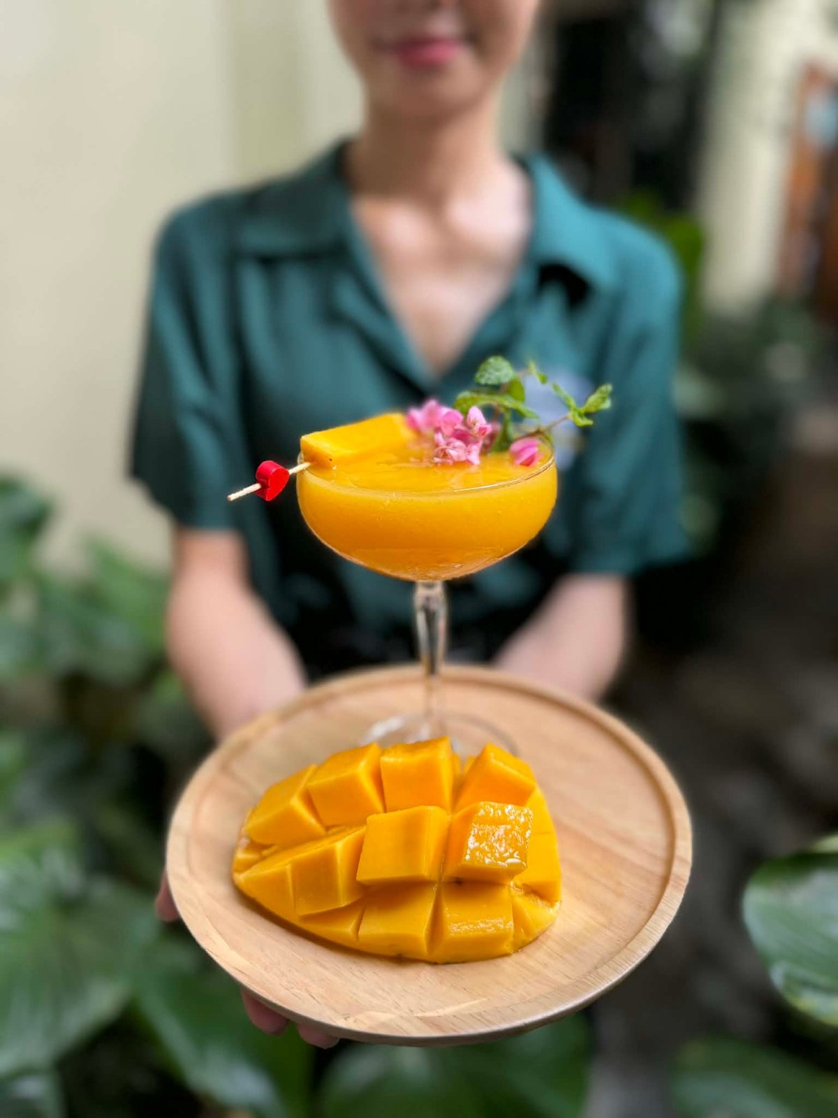 Bartender in teal shirt holding wooden plate with sliced mango and yellow cocktail garnished with pink flower and cherry