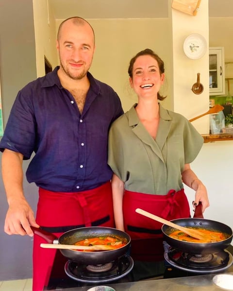 A man in a navy shirt and red apron and a woman in a sage green shirt and red apron smile while cooking together at a stovetop in a modern kitchen